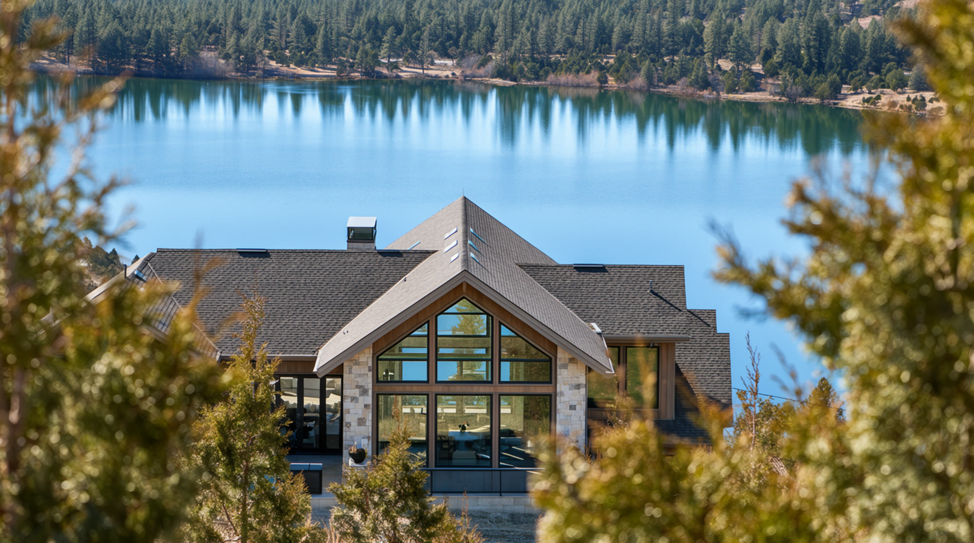 A steep gable and a tall wall of glass in stone and timber, framed by pines with a lake behind.